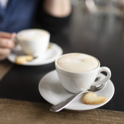 Eine Person sitzt vor einer Kaffeetasse. Davor steht eine zweite Tasse mit einem Herz-Biskuit.