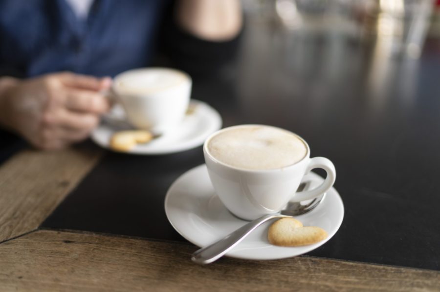 Eine Person sitzt vor einer Kaffeetasse. Davor steht eine zweite Tasse mit einem Herz-Biskuit.